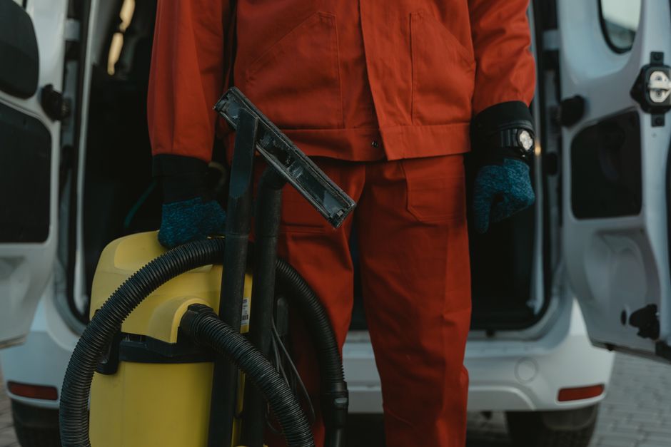 A professional cleaner dressed in a red uniform and gloves standing in front of an open vehicle door, holding a yellow vacuum and cleaning tools. The background shows the interior of their cleaning va