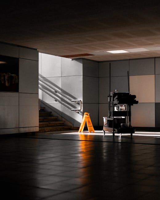 A dimly lit indoor space featuring a staircase with metal handrails, partially illuminated by natural light casting shadows on the tiled floor. To the right of the staircase, a cleaning cart equipped 