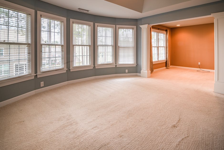 A spacious, well-lit residential living room with a large bay of windows fitted with white blinds, allowing natural light to illuminate the beige plush carpeted floor. The walls are painted in soft pa