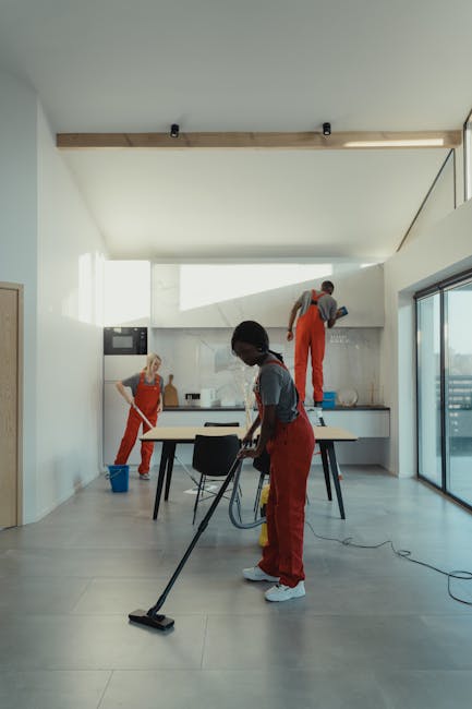 Two professional cleaners from Cleaners Crystal Palace are engaged in a domestic cleaning task inside a modern living room. One cleaner is kneeling on the light grey tiled floor, holding a vacuum clea